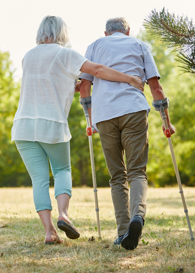 Two elderly individuals walking together, one using a cane and the other with crutches, in a park setting.